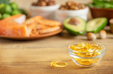 A variety of nutrient rich foods are arranged on a wooden surface with soft, natural light. The foreground features a glass bowl filled with golden fish oil capsules. In the blurred background are foods such as salmon, avocado, and various nuts and seeds.