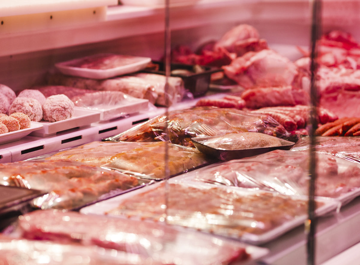 A display case in a shop contains different cuts and preparations of meat. Some are wrapped in clear plastic on trays and some are arranged on serving dishes. Meatballs and sausages are visible