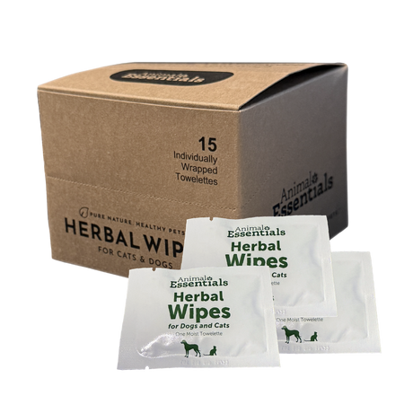 A brown box labeled Animal Essentials | Herbal Wipes (15 count box) sits with three individual white towelette packets displayed in front. Designed for natural grooming, the box notes it contains 15 individually wrapped herbal pet wipes for cats and dogs.