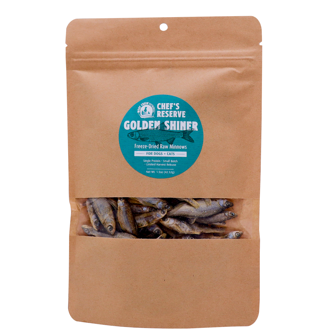 A brown resealable pouch with a clear window displays freeze-dried minnows inside. The label reads Woof Creek Golden Shiner Minnows | 1.5oz Pouch for Dogs + Cats. These nutritious morsels are perfect as dog treats or cat treats and make an excellent healthy reward for training.