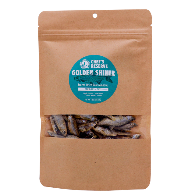 A brown resealable pouch with a clear window displays freeze-dried minnows inside. The label reads Woof Creek Golden Shiner Minnows | 1.5oz Pouch for Dogs + Cats. These nutritious morsels are perfect as dog treats or cat treats and make an excellent healthy reward for training.