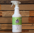 A white spray bottle of Kin+Kind Flea/Tick Repel Spray for Dogs Only | Lavender, a lavender-scented plant-based pet spray, sits on a wooden surface with a wood panel background. The green and white label has purple and black text.