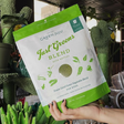 A hand holds a bag of Green Juju Just Greens - Freeze Dried Green Vegetable Blend for dogs, featuring organic nettles, with green plants and store shelves in the background.